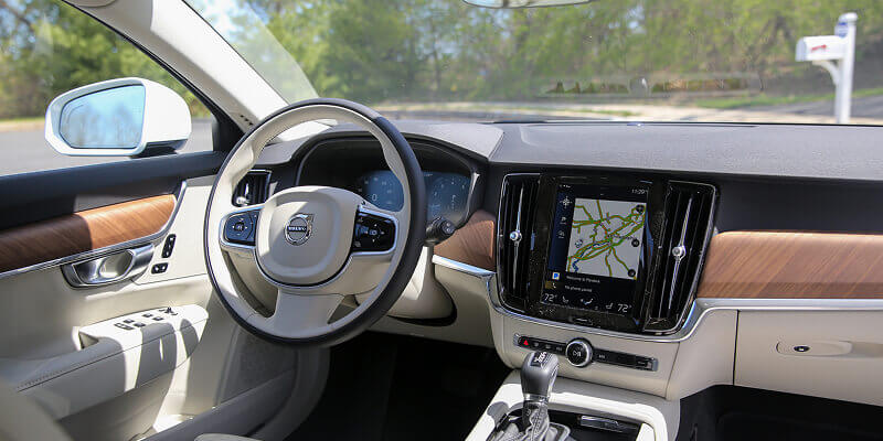 The car dashboard features a steering wheel with a logo, a touchscreen displaying a map, and wood accents. The car is parked in a sunny area with greenery visible outside.