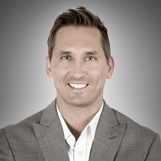 A man smiles while wearing a gray suit with a white dress shirt. The background is a neutral gray gradient.