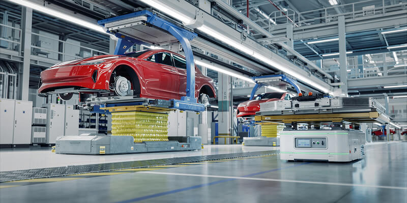 Red car frames are elevated on blue lifts inside a modern, spacious, and well-lit automotive assembly line, surrounded by machinery, white cabinets, and overhead lighting stretching across the factory floor.