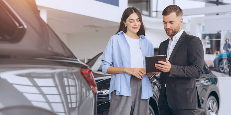 A man and woman stand in a car showroom. The man holds a tablet they both examine, surrounded by several cars, emphasizing a business or sales discussion.