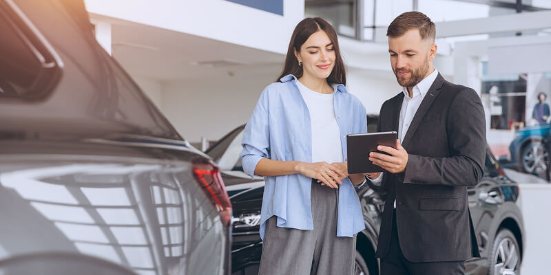 A man and woman stand in a car showroom. The man holds a tablet they both examine, surrounded by several cars, emphasizing a business or sales discussion.