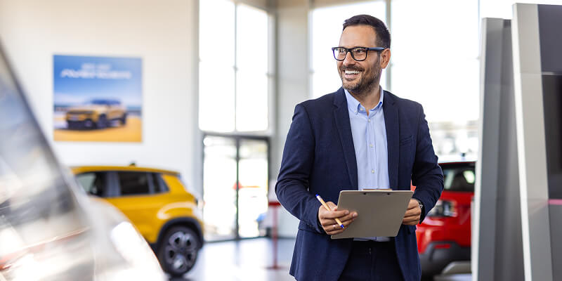 A man in a suit holds a clipboard and smiles, standing in a car showroom. He is near several vehicles, with large windows providing bright natural light.