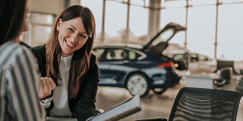 A woman smiles while holding a document, engaging with another person. They are seated in a car dealership, with a blue car visible in the background, trunk open.