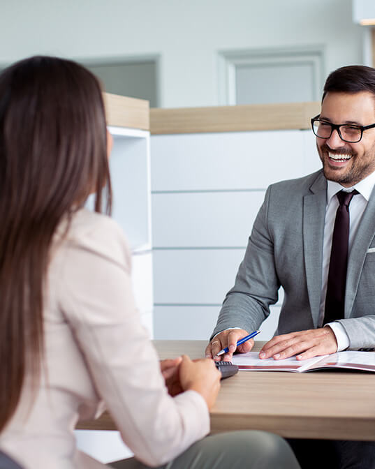 A man in a suit smiles while discussing something with a woman across a table, in a modern, well-lit office setting.