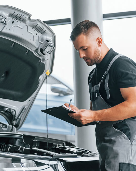 A mechanic inspects and writes on a clipboard next to an open car hood in a garage, surrounded by large windows and bright natural light.