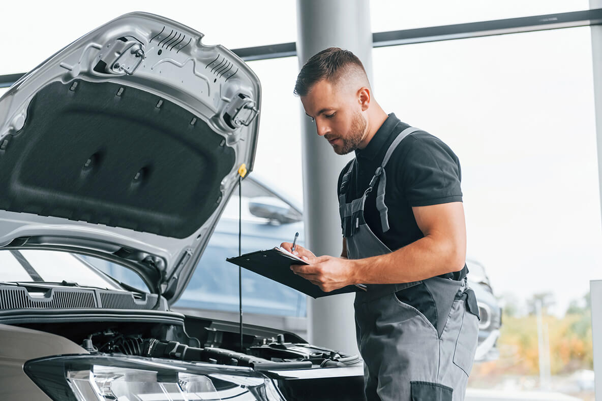 A mechanic inspects a car engine, writing on a clipboard. The car hood is open, and they are in a bright, modern garage with large windows.