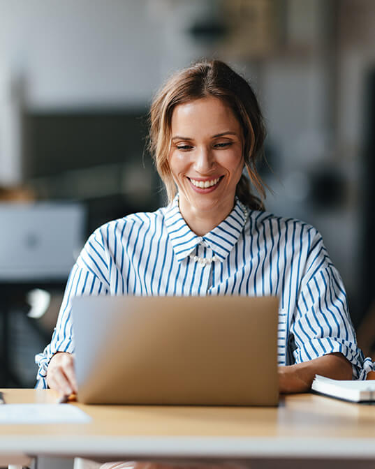 A person is smiling while using a laptop at a desk in a bright, modern office setting.