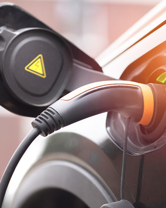 Charging plug inserted into an electric vehicle, with a visible yellow warning symbol on a nearby cap, set against a blurred outdoor background.