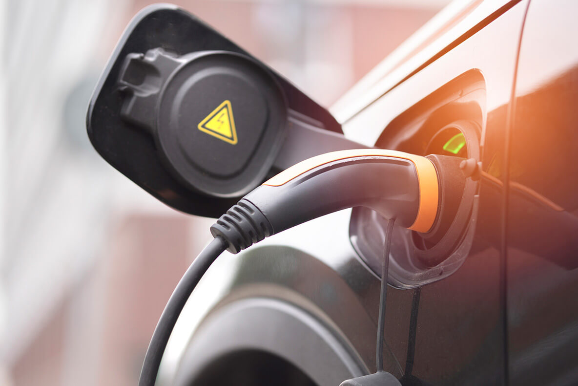 Electric vehicle charging cable plugged into a car, with a black cap open and a yellow triangular warning symbol visible, in an outdoor setting.