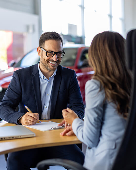 A man in glasses, smiling and writing on a notepad, sits at a table across from a woman in a business setting with a red car in the background.