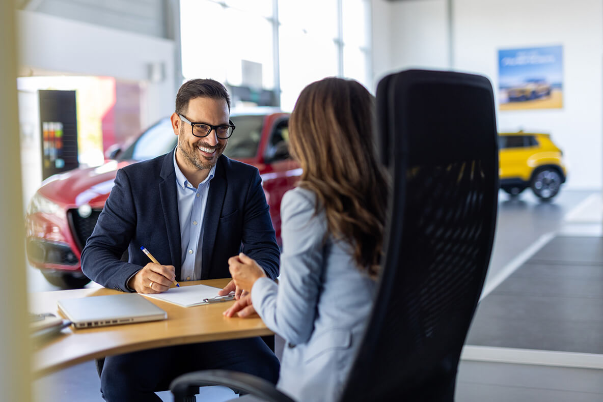 Man writing with pen on clipboard, smiling at woman seated across a table in a car dealership. Red and yellow cars are displayed in the background beside large windows.