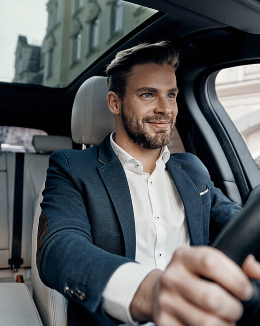 A man in a suit drives a car, smiling confidently, with urban buildings visible through the windows.