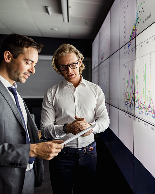 Two men discuss, holding a paper, in front of a large screen displaying colorful graphs and charts. They are in a modern, professional office setting.