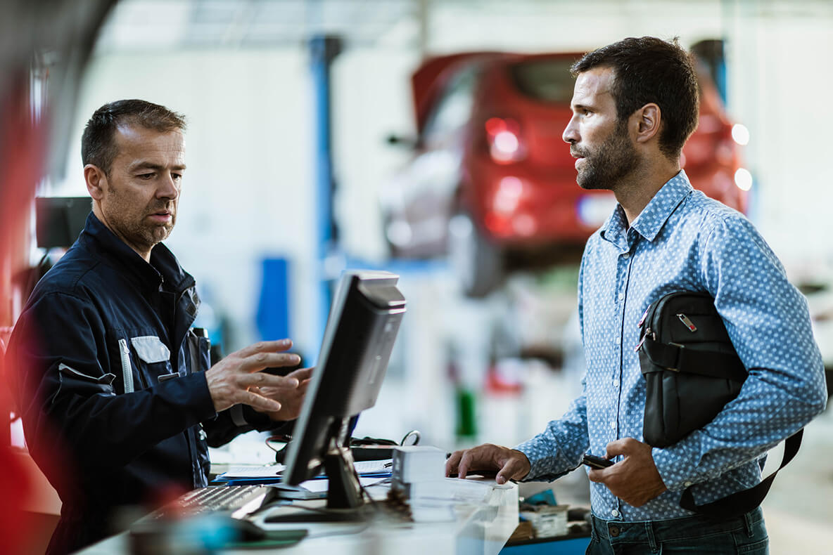 A person in a mechanic's uniform gestures while discussing with another person holding a bag, standing at a counter in an auto repair shop, with a red car on a lift in the background.
