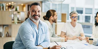 A bearded man in a light blue shirt smiles and holds a pen, sitting at a desk. Behind him, three people are engaged in discussion in a bright office setting.