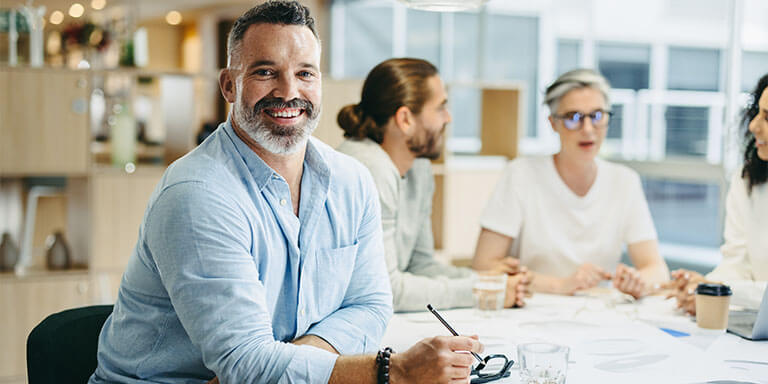 A person smiles and holds a pen while sitting at a table. In the background, three people engage in conversation, creating a collaborative office setting.