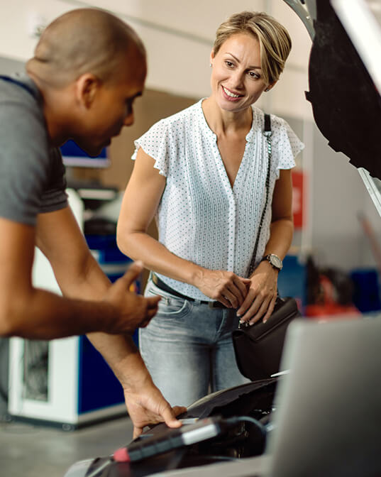 Mechanic explains car engine details to a woman, who is attentively listening, in a garage setting with vehicles and equipment in the background.