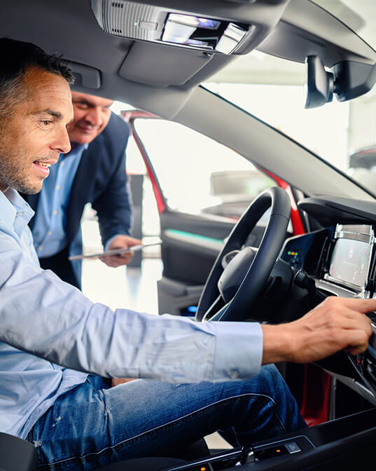 A man sits in a car's driver's seat, adjusting the dashboard controls, while another man observes with a clipboard. The car is in a showroom setting.