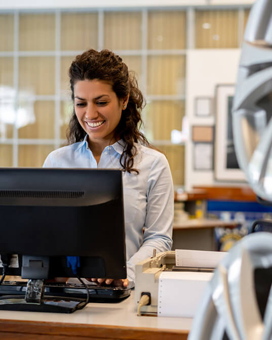 A woman in a light blue shirt smiles while typing on a computer keyboard. She's in an office with a grid-patterned window and framed pictures in the background.