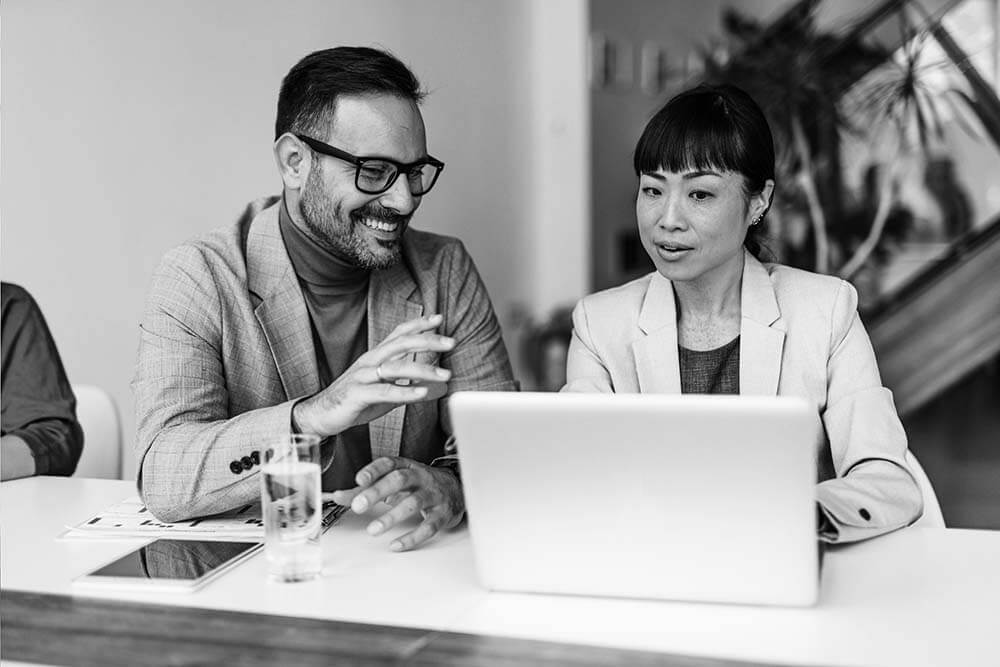 Two people in business attire sit at a table, discussing something on a laptop. A tablet and a glass of water are on the table, with plants in the background.