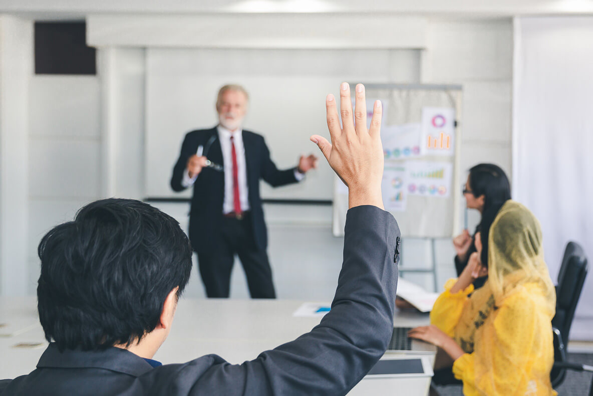 A person raises their hand, facing an older man in a suit giving a presentation. The room contains a flipchart with graphs and several seated participants.