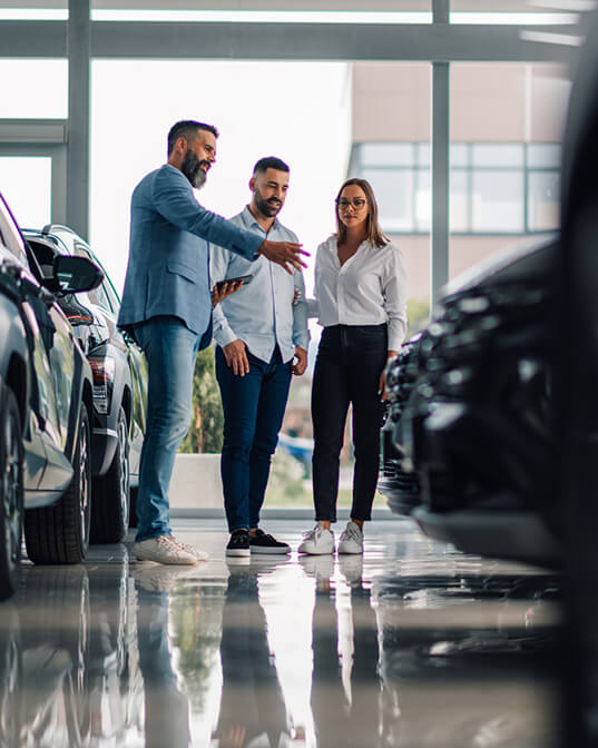 Three people stand in a car dealership, discussing vehicles parked on a shiny showroom floor surrounded by large windows.