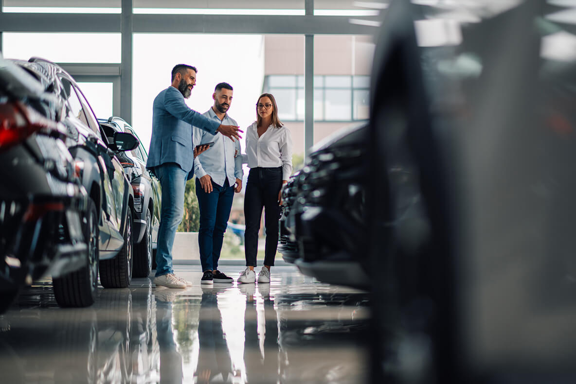 Three people stand in a car dealership, discussing vehicles parked on a shiny showroom floor surrounded by large windows.