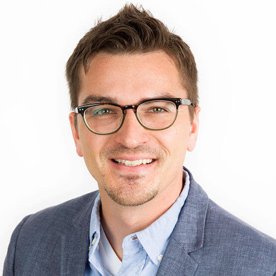 A smiling person with glasses and short hair wears a blue suit jacket and light shirt, against a plain white background.