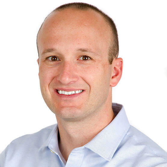 A person with short hair smiles, wearing a light blue shirt against a plain white background.