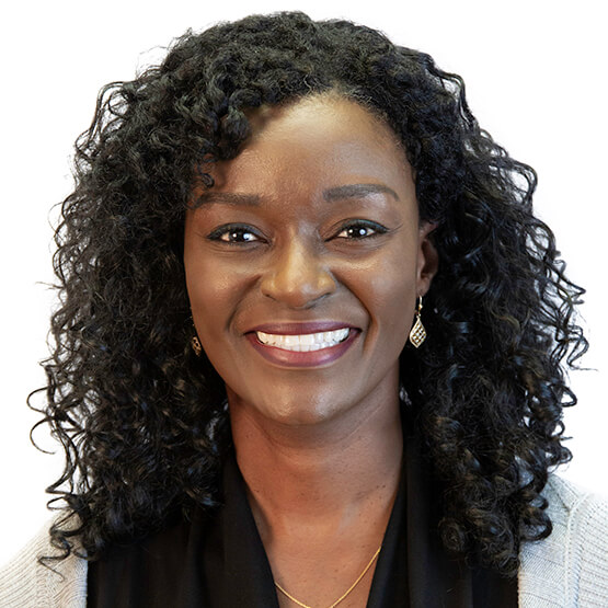 A person with curly hair is smiling directly at the camera, wearing earrings and a dark top, set against a plain white background.