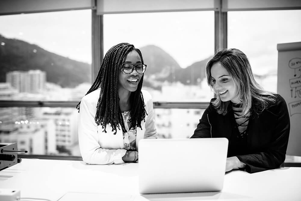 Two women sit at a table, smiling while looking at a laptop. They are in a bright office with large windows and cityscape in the background.