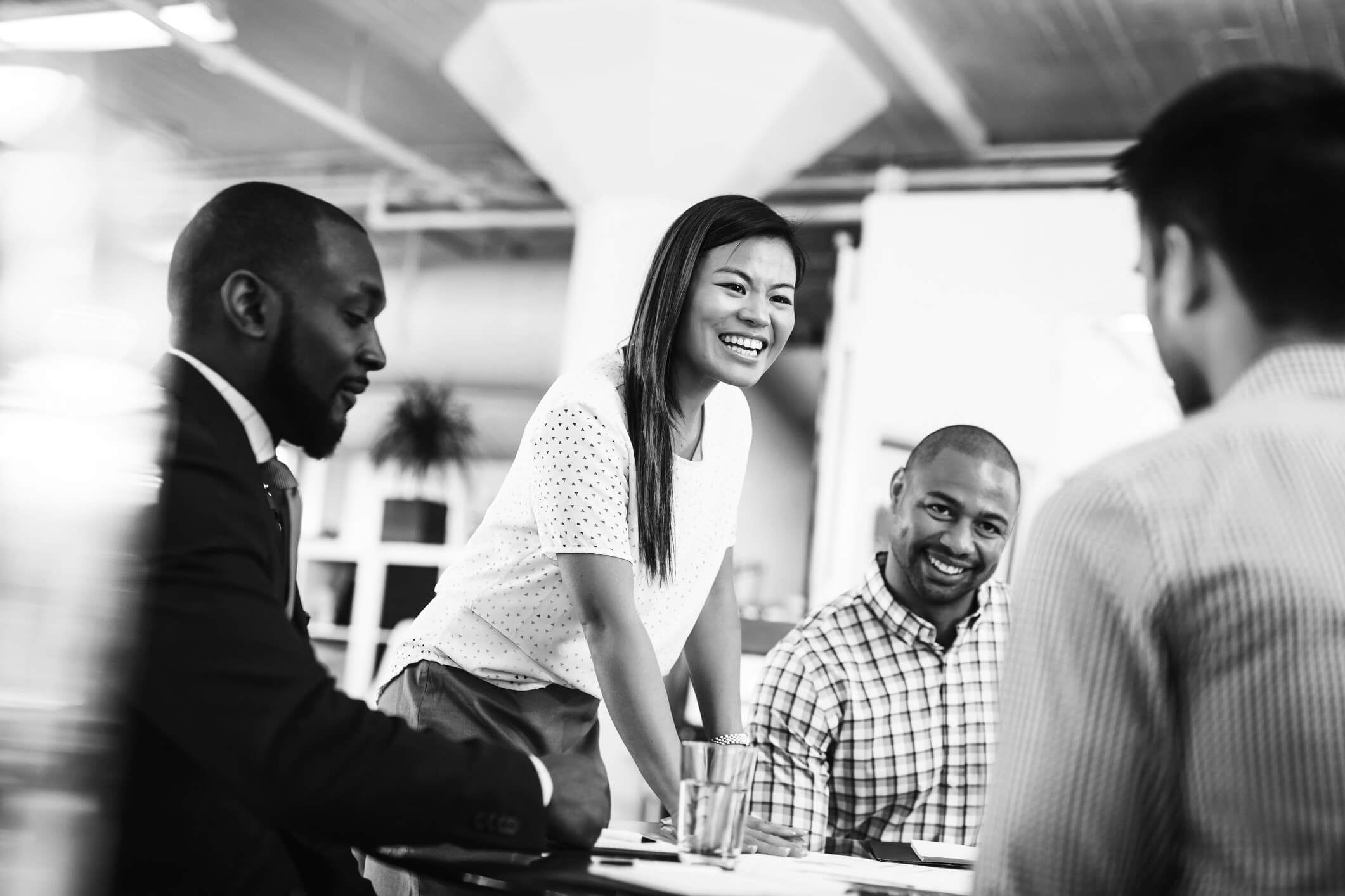 Four people gather around a table in an office setting. A woman smiles while speaking, and others appear engaged. Glasses, papers, and a plant are in the background.
