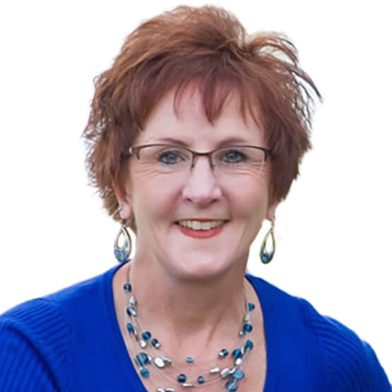 A person with short reddish hair and glasses smiles, wearing blue attire and beaded jewelry, set against a plain white background.