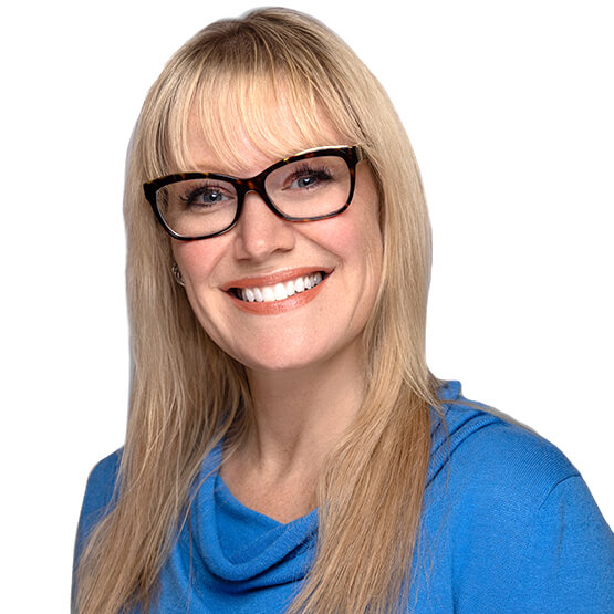 Person with long blonde hair and bangs, wearing tortoiseshell glasses and a blue cowl-neck top, smiles directly at the camera against a plain white background.