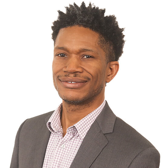 Person smiles subtly, head turned slightly to the left, wearing a gray pinstripe blazer over a pink checkered shirt; cropped curly hair and light stubble against a plain white studio background.