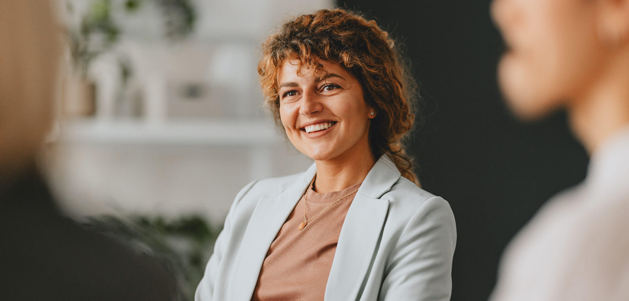 Young woman with curly hair smiles warmly while listening and engaging in conversation in a modern office; blurred colleagues in the foreground suggest a meeting setting.