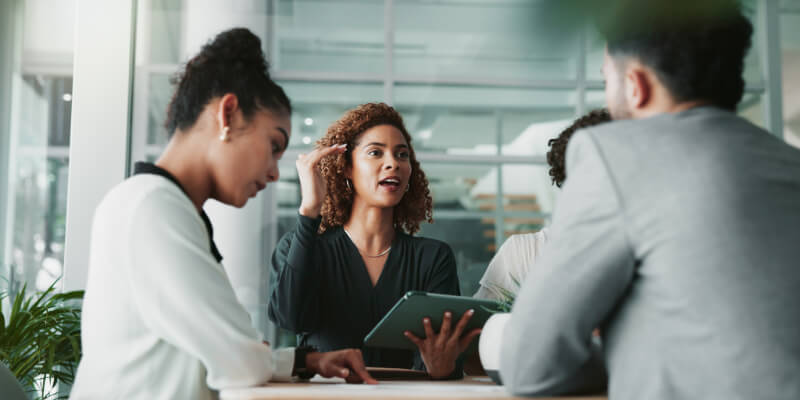 Central woman gestures while holding a tablet, addressing three colleagues seated around a conference table in a bright, glass-walled modern office.