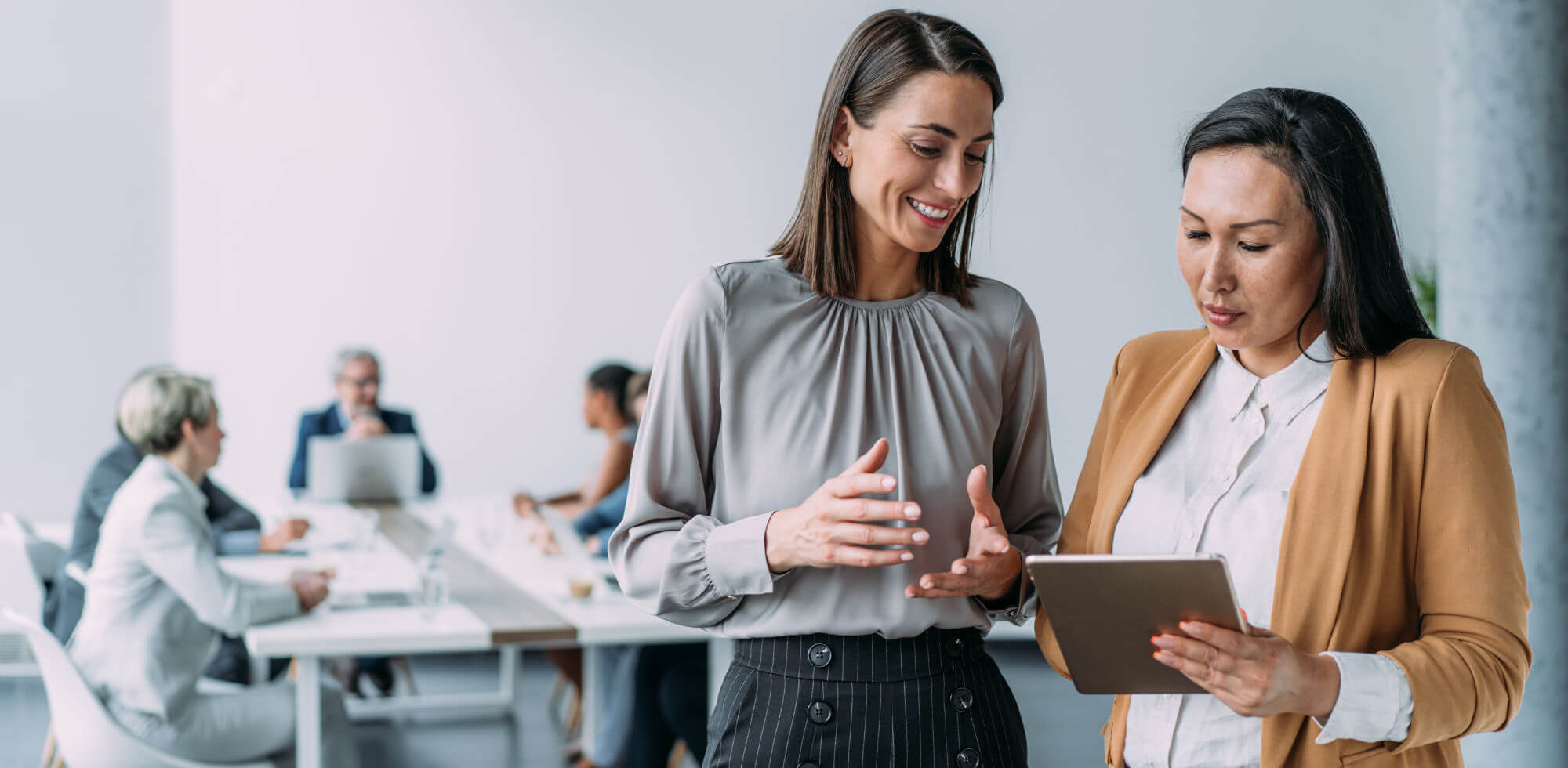 Two professional women review a tablet; one gestures and smiles while the other reads, standing beside a conference table where colleagues meet in a bright office.