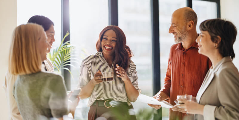 A smiling woman holds a coffee mug and laughs while speaking; four coworkers listen, some holding drinks and papers, gathered in a sunlit modern office near large windows.