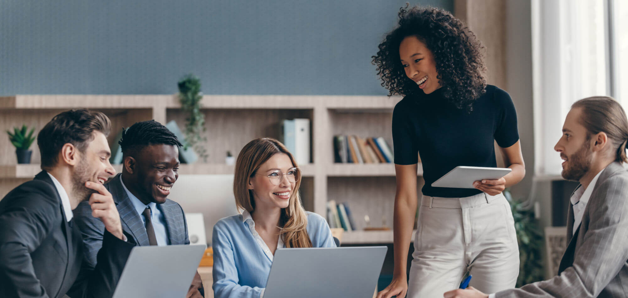 A group of five colleagues (three men and two women) smile and converse while viewing laptops and a tablet in a modern office with bookshelves, plants, and a window-lit workspace.