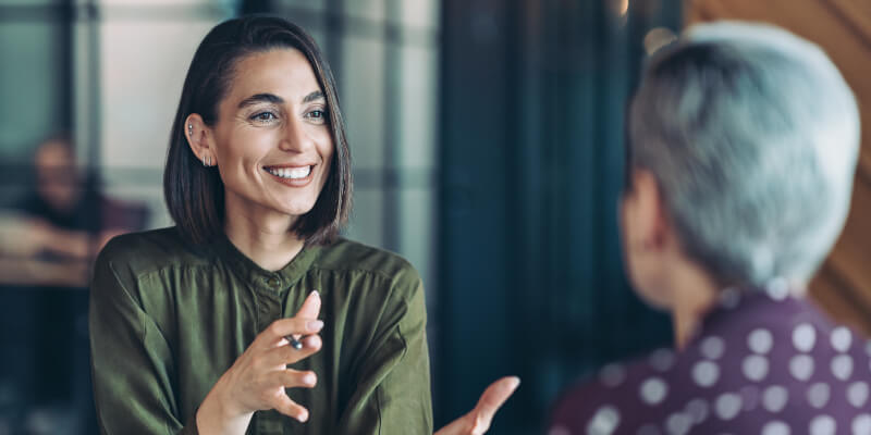 Woman in a green blouse smiles and gestures while speaking to another seated person with short gray hair, in a softly lit modern office with a blurred background.