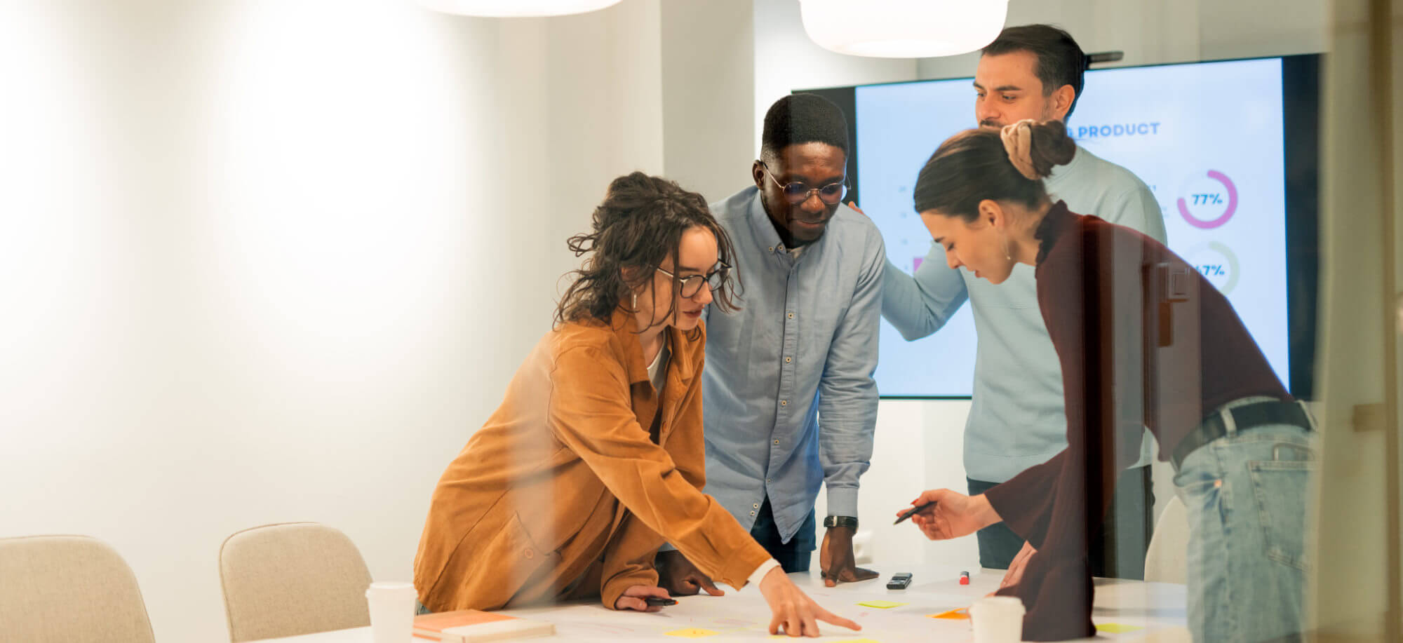 Four colleagues leaning over a table, pointing at papers and sticky notes while discussing; glass-walled conference room with a wall monitor displaying circular charts.