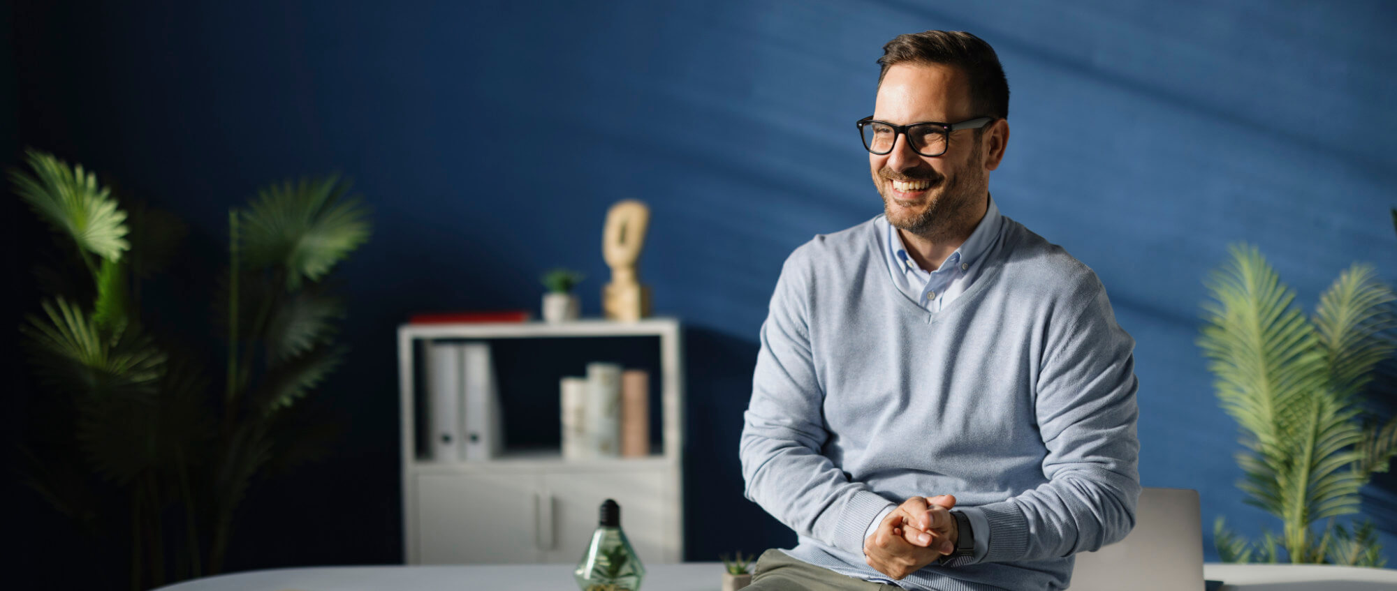 Smiling man wearing glasses and a light-blue sweater sits on a desk edge, hands clasped, looking right; office setting with a blue wall, potted plants, white shelf and decorative objects.