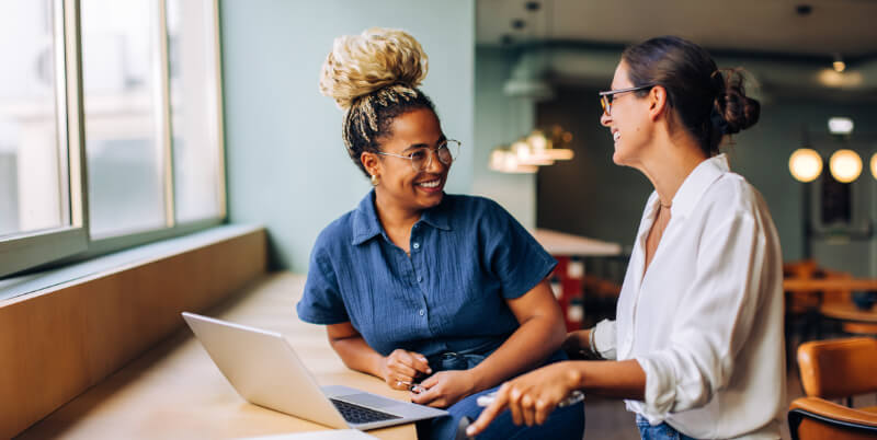 Two women sit at a window-side counter with an open laptop, smiling and talking; one gestures while the other listens, in a bright café-like coworking space with pendant lights.