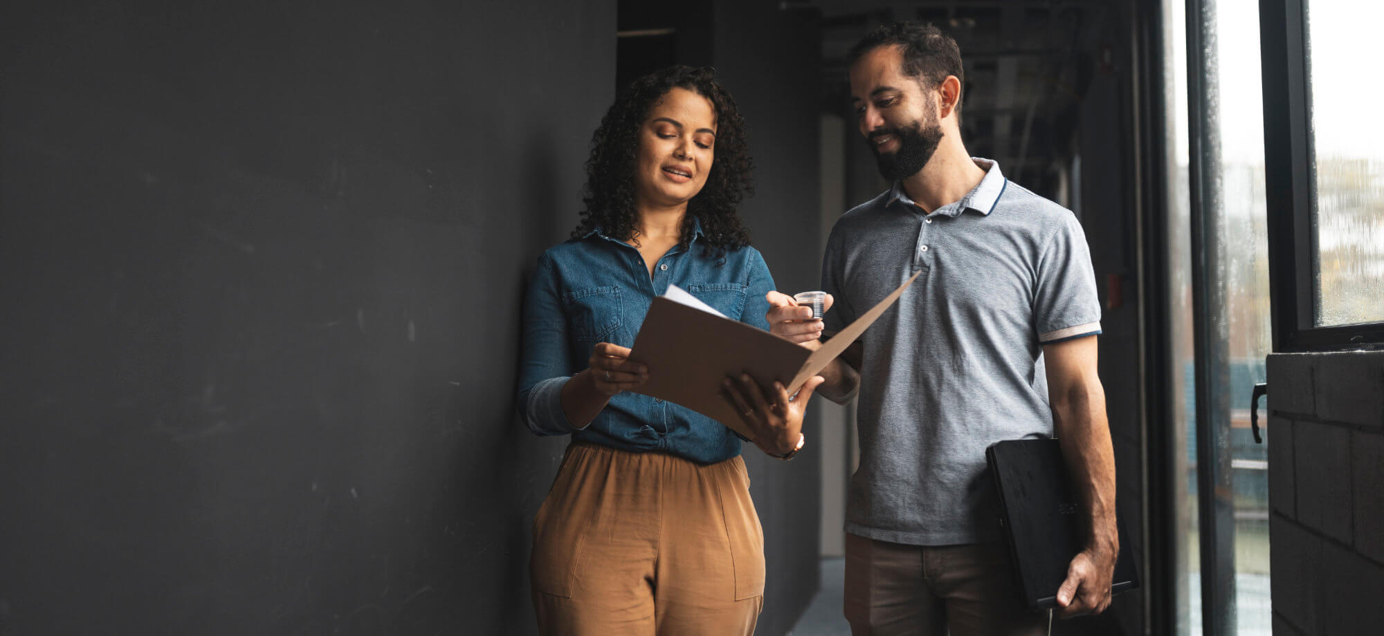 Woman holding an open folder shows documents to a man who points and holds a coffee cup and laptop, standing in a dim office corridor by a window.