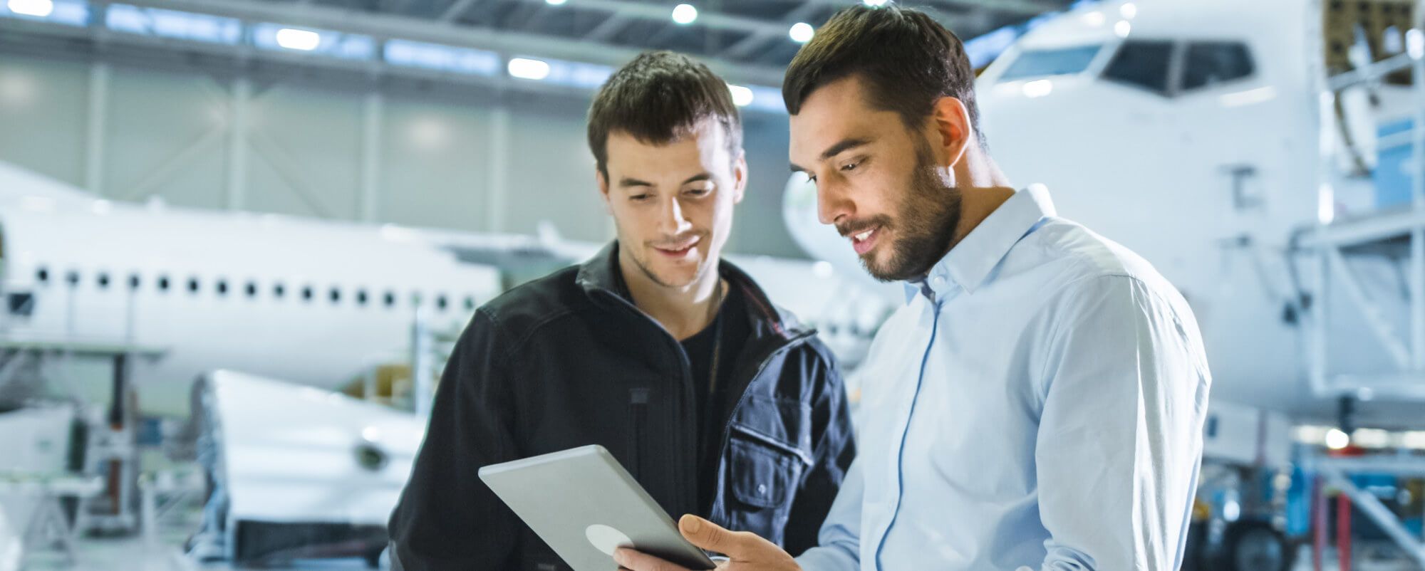 Two men study a tablet together, discussing results, standing in a maintenance hangar with a partially visible commercial airplane and equipment blurred behind them.