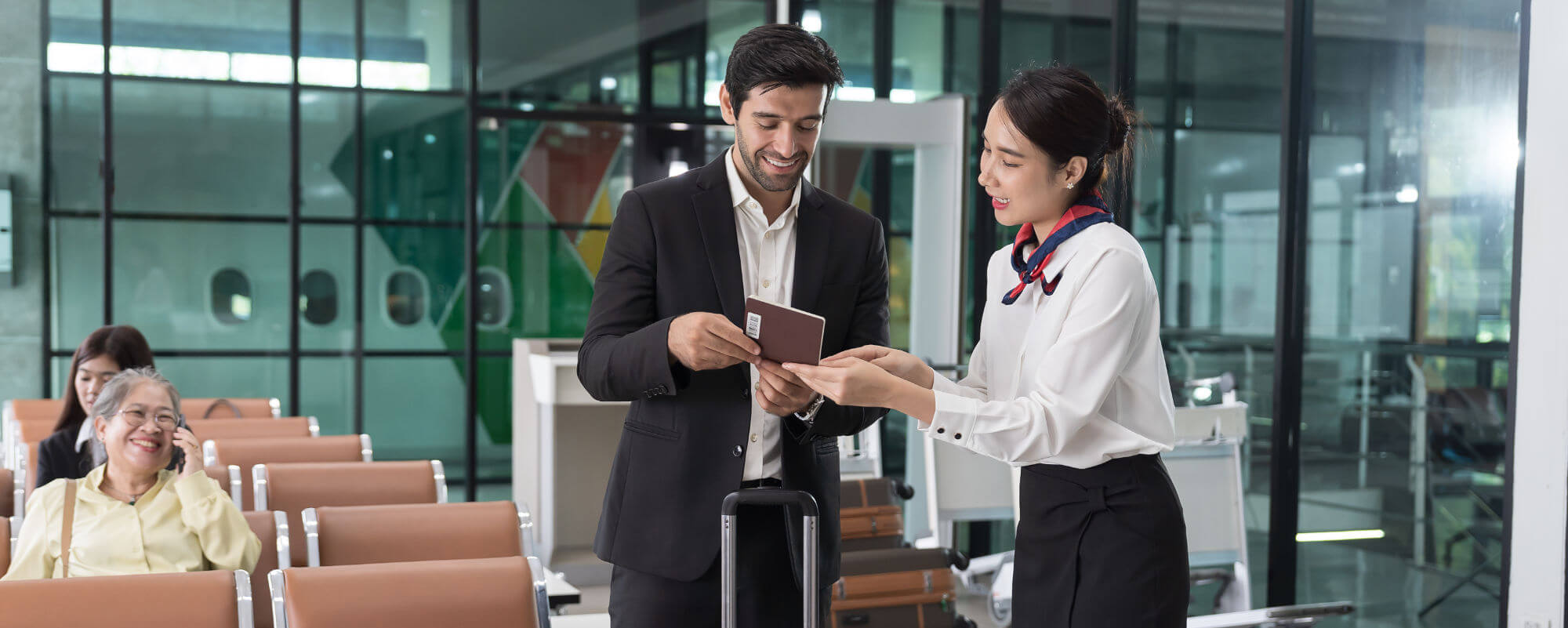 Male traveler hands a passport to a female airline attendant while smiling; they stand beside a carry-on in an airport departure lounge with rows of seats and glass partitions.