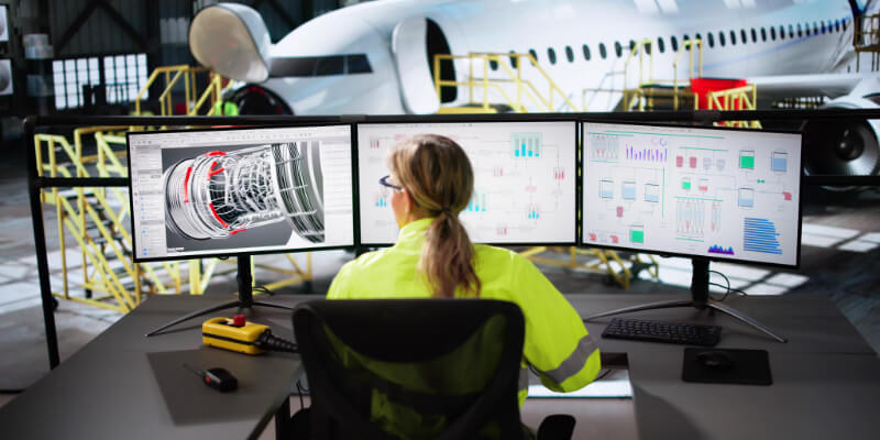 A technician sits at three monitors analyzing a jet-engine 3D model and flight-data graphs, inside an aircraft hangar with a commercial airliner and maintenance platforms behind.
