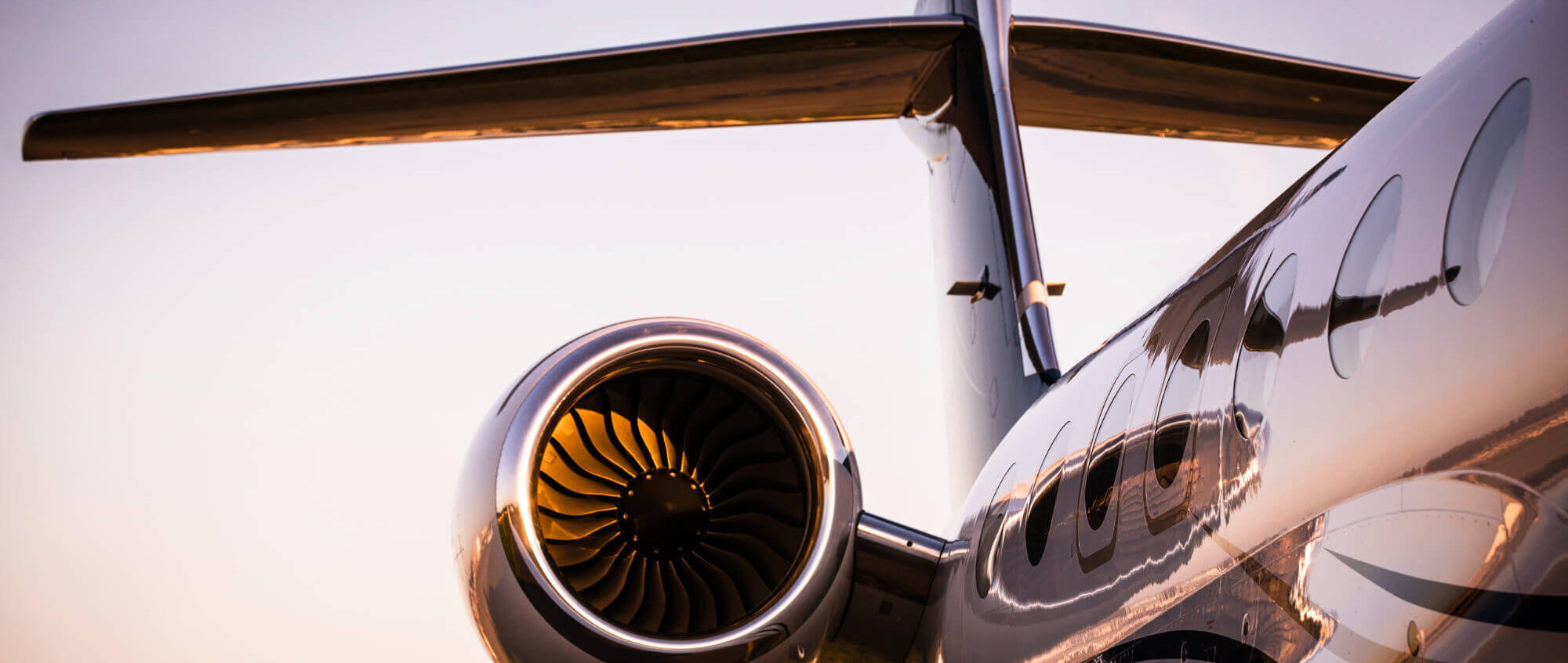 Jet turbofan gleams in warm sunset light, sitting idle; mounted on a sleek private jet fuselage with a T-tail and oval windows reflecting runway and sky.