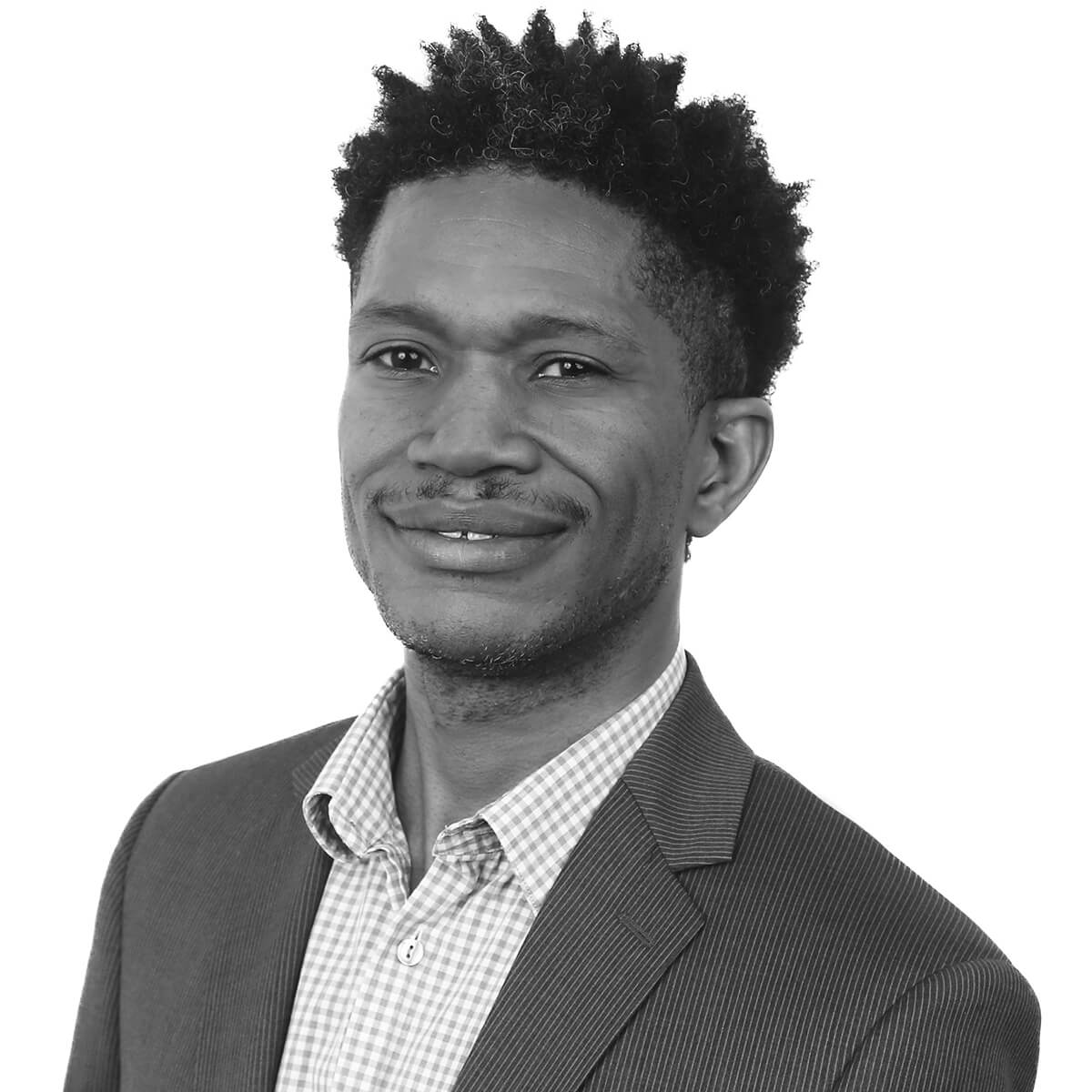 Man smiles slightly while facing the camera, wearing a pinstripe suit jacket and checked shirt; short curly hair and trimmed facial hair; head-and-shoulders portrait against a plain white background.